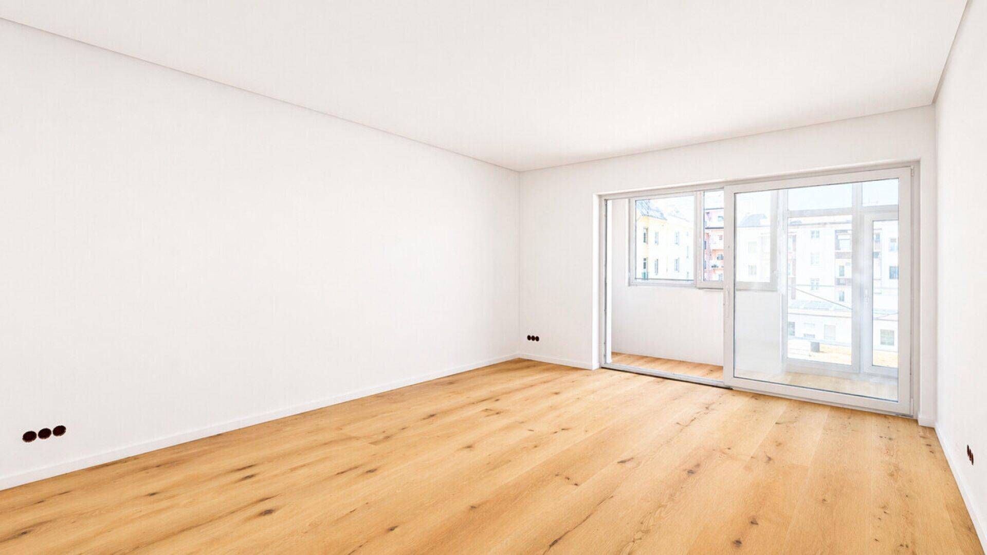 Helles, leeres Zimmer mit neuem Holzfußboden und Zugang zu einem Balkon.