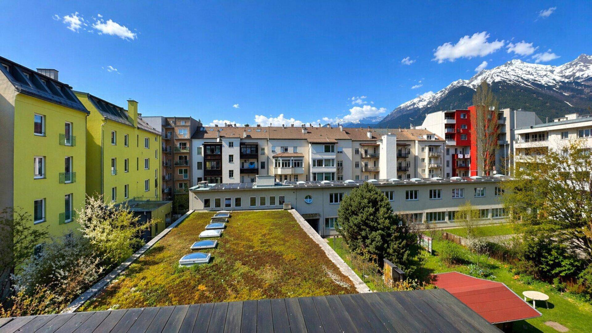 Panoramablick auf die umliegenden Gebäude, grüne Dächer und schneebedeckte Berge im Hintergrund.