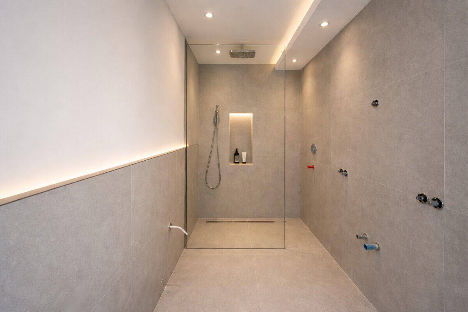 Modernes Badezimmer mit begehbarer Dusche, grauen Fliesen und indirekter Beleuchtung.
