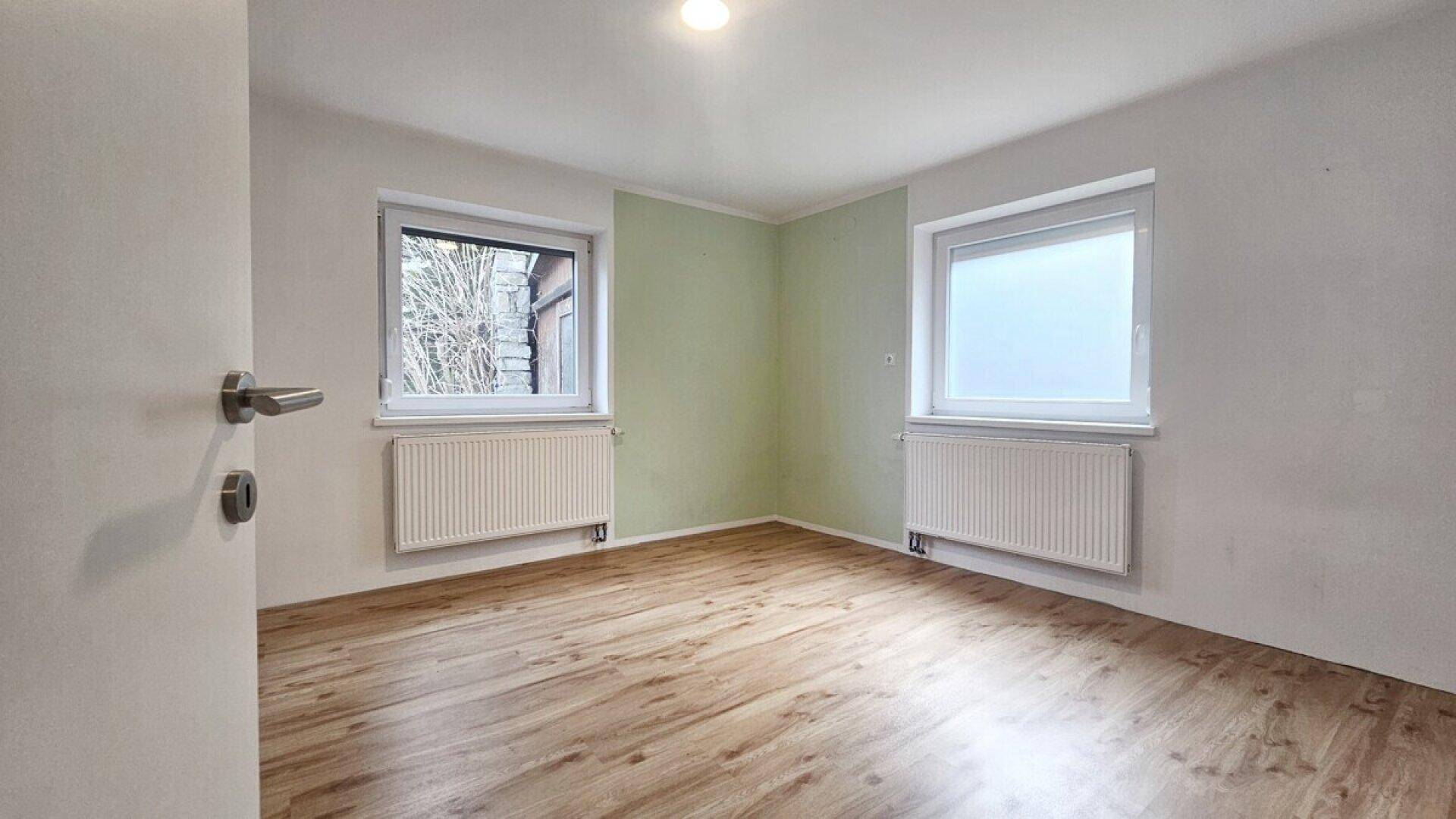 Helles Schlafzimmer mit Laminatboden, einer grünen Akzentwand und zwei Fenstern.