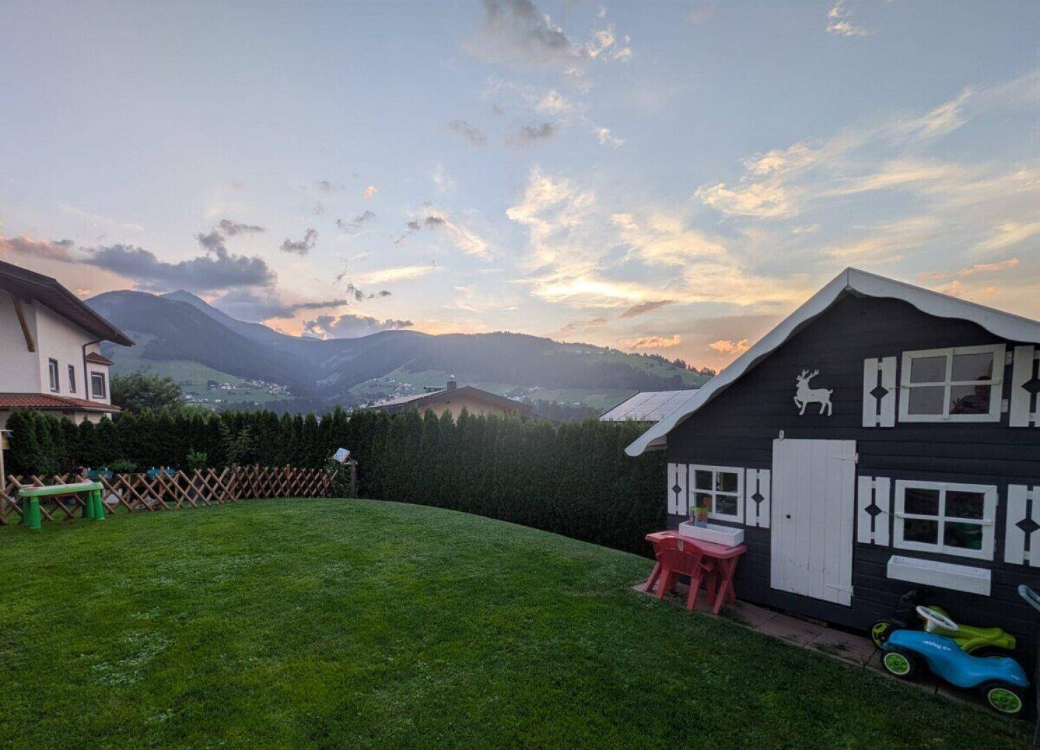 Abendstimmung im Garten mit Spielhaus, Rasenfläche und Blick auf die umliegenden Berge.