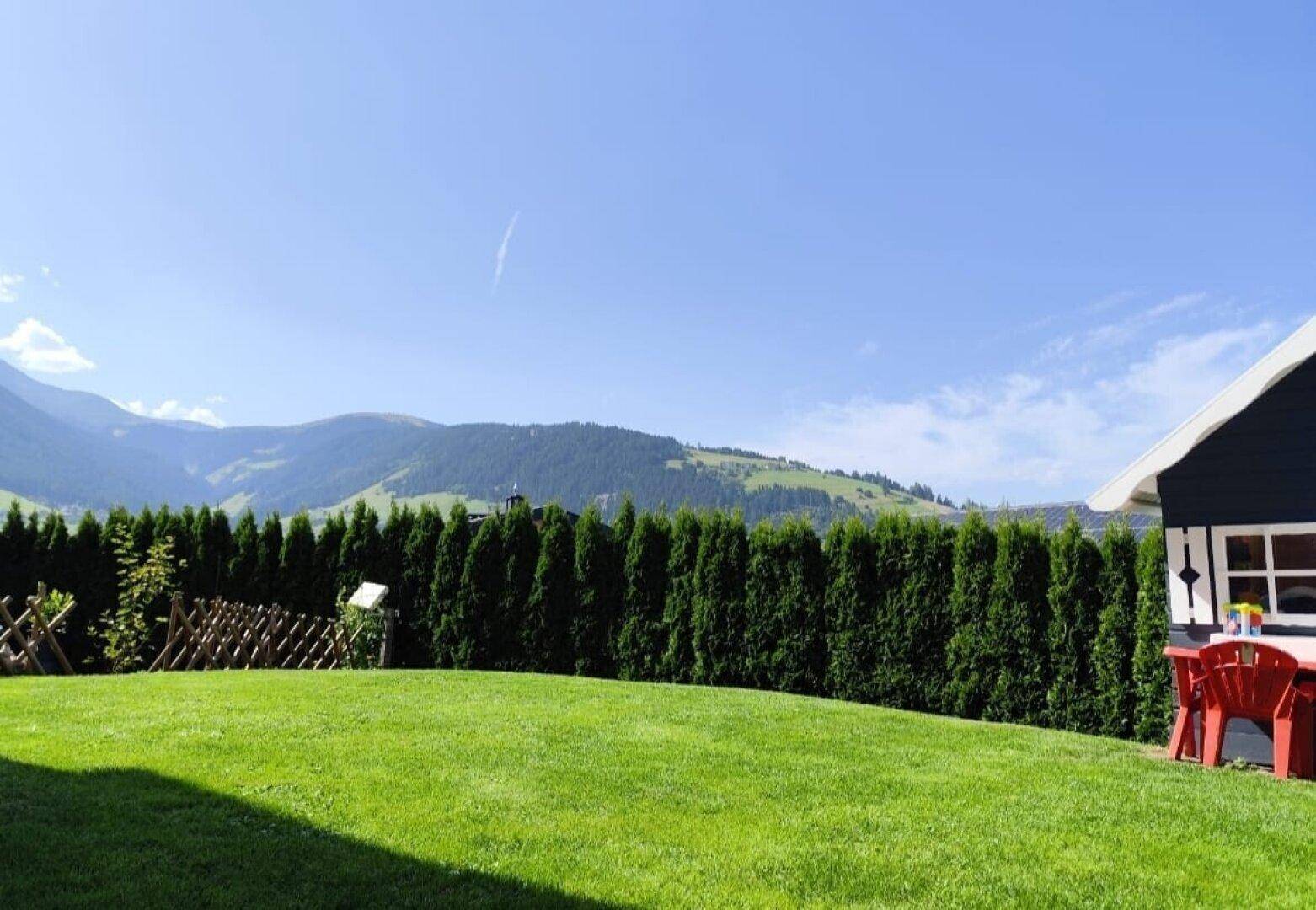 Sonniger Garten mit gepflegtem Rasen, hoher Hecke und malerischem Bergblick.