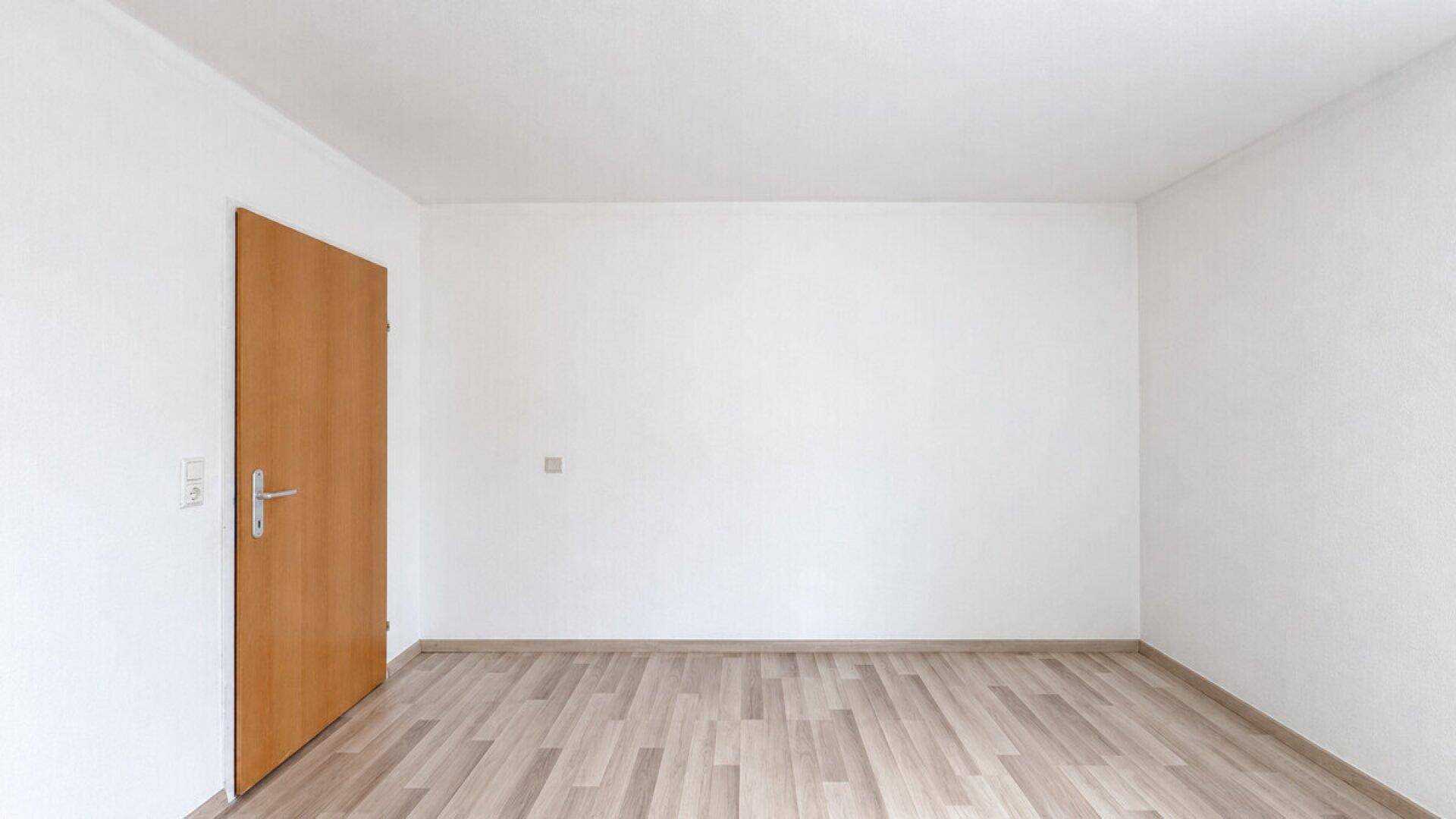 Helles, leeres Zimmer mit Laminatboden und einer Holztür, ideal als Schlaf- oder Arbeitszimmer.