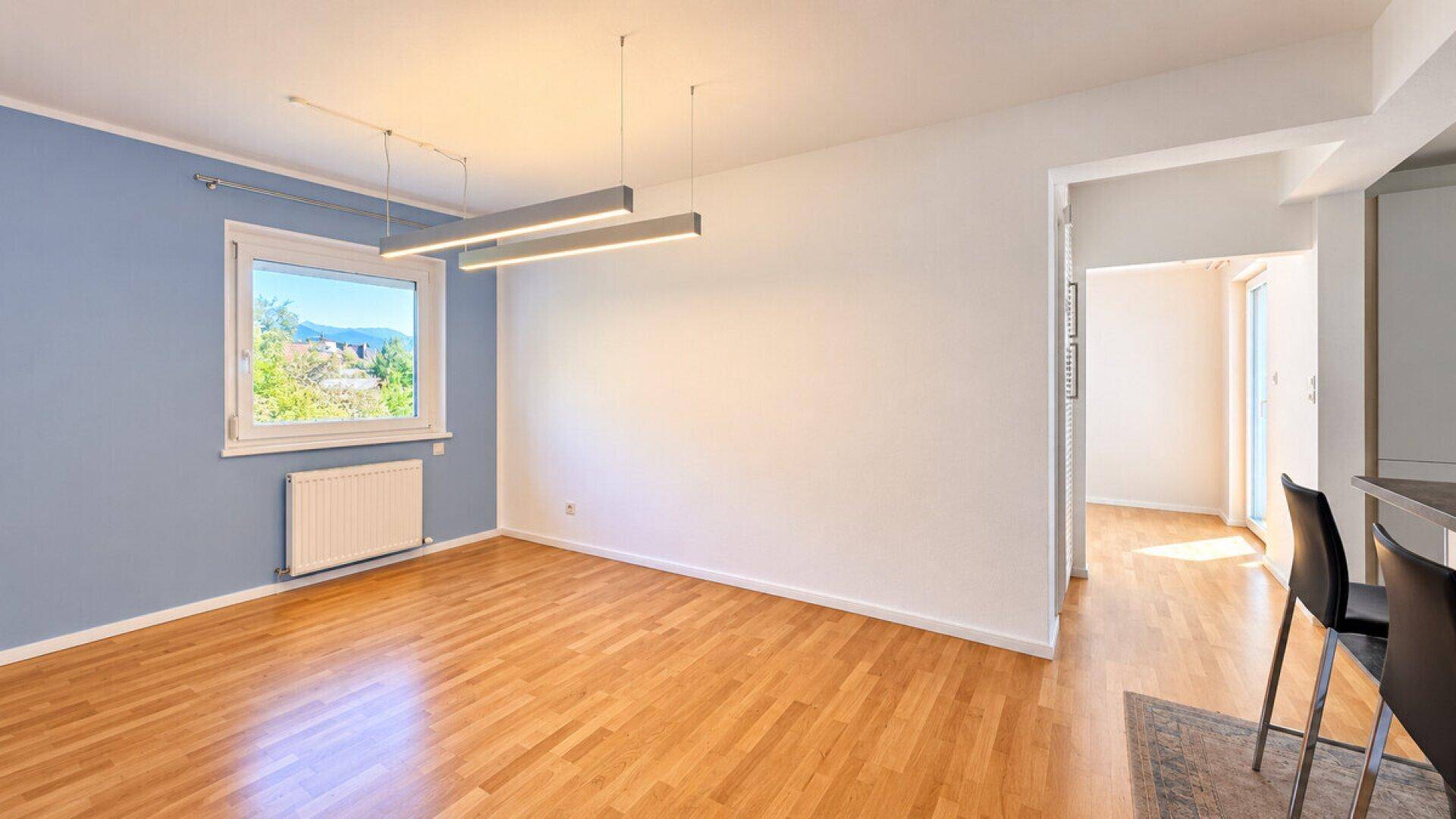 Heller Wohnraum mit Parkettboden, blauer Akzentwand und großem Fenster mit Ausblick.