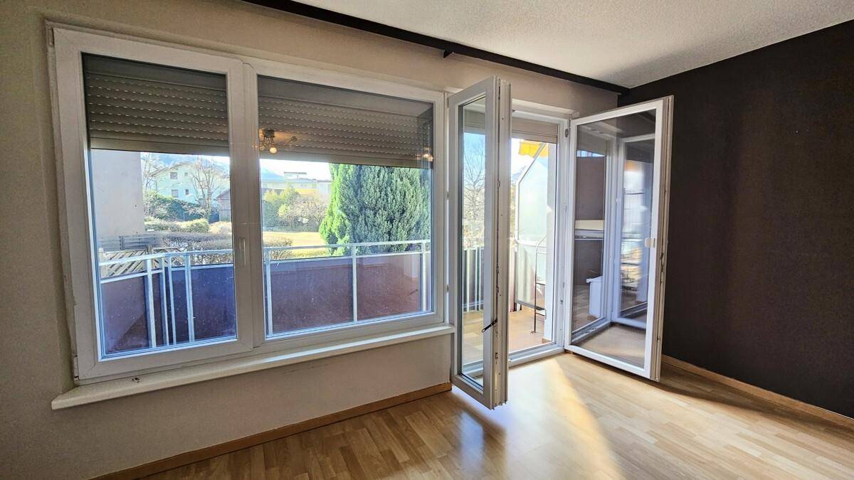 Heller Raum mit Holzboden, großen Fenstern und Zugang zu einem Balkon mit grüner Aussicht.