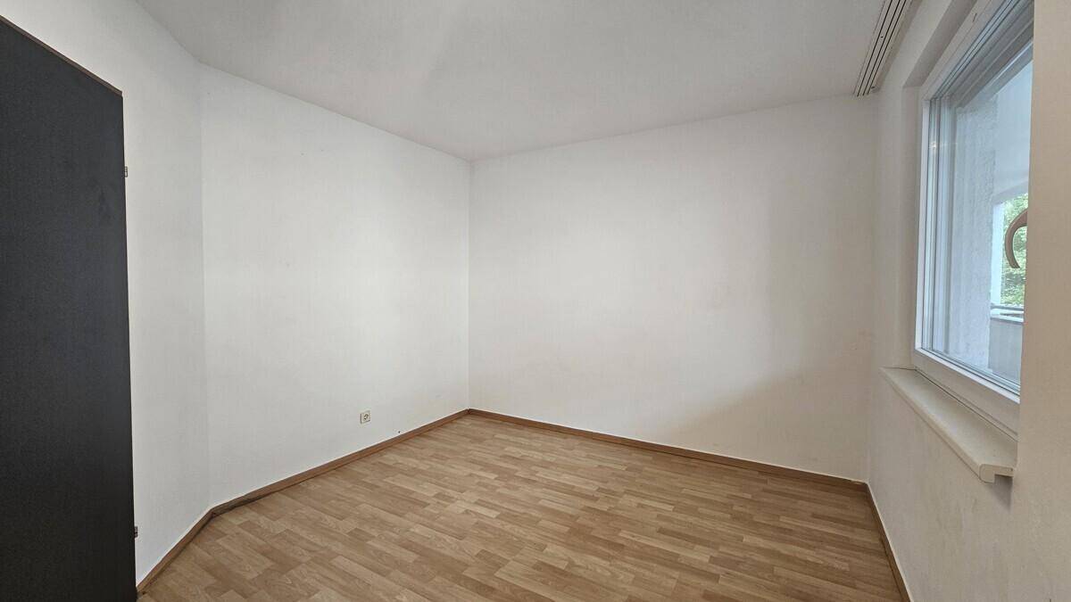 Helles, leeres Zimmer mit weißen Wänden, Holzboden und einem Fenster.