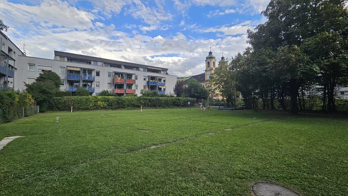Grüne Rasenfläche vor einem Wohngebäude mit Balkonen und einer Kirche im Hintergrund.