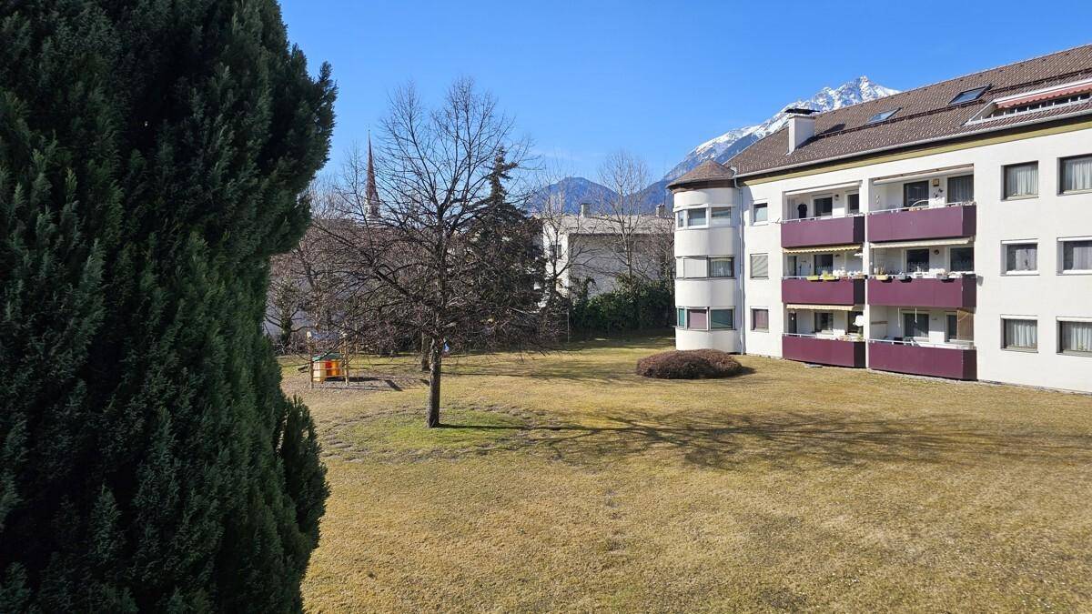 Außenansicht eines Wohngebäudes mit großer Rasenfläche, Bäumen und einem Spielplatz im Vordergrund.