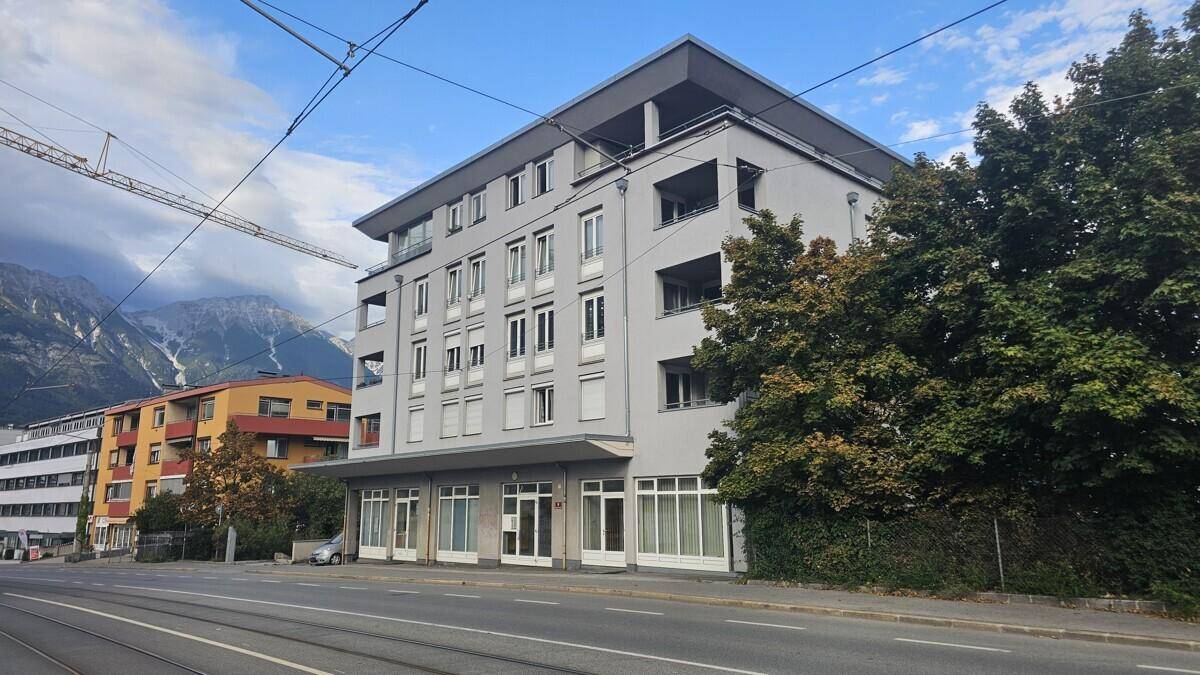 Mehrfamilienhaus an einer Straße mit Bergen im Hintergrund unter blauem Himmel.