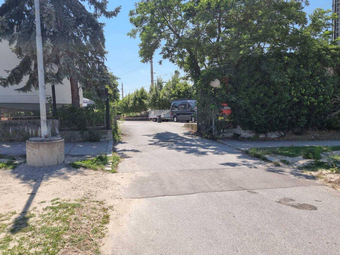 Zufahrt zu einem Grundstück mit Bäumen und einem Fahrzeug im Hintergrund an einem sonnigen Tag.