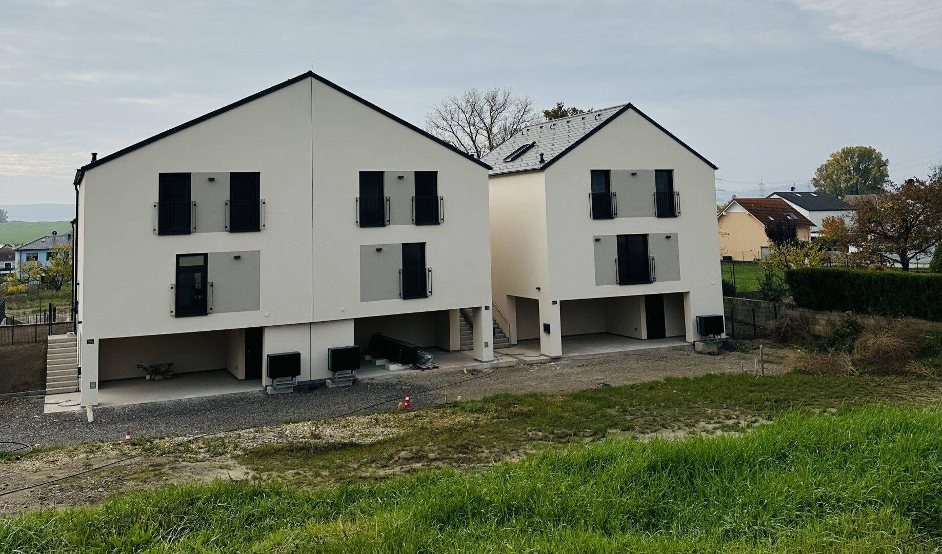 Zwei moderne Reihenhäuser mit weißer Fassade und dunklen Fensterläden in ländlicher Lage.