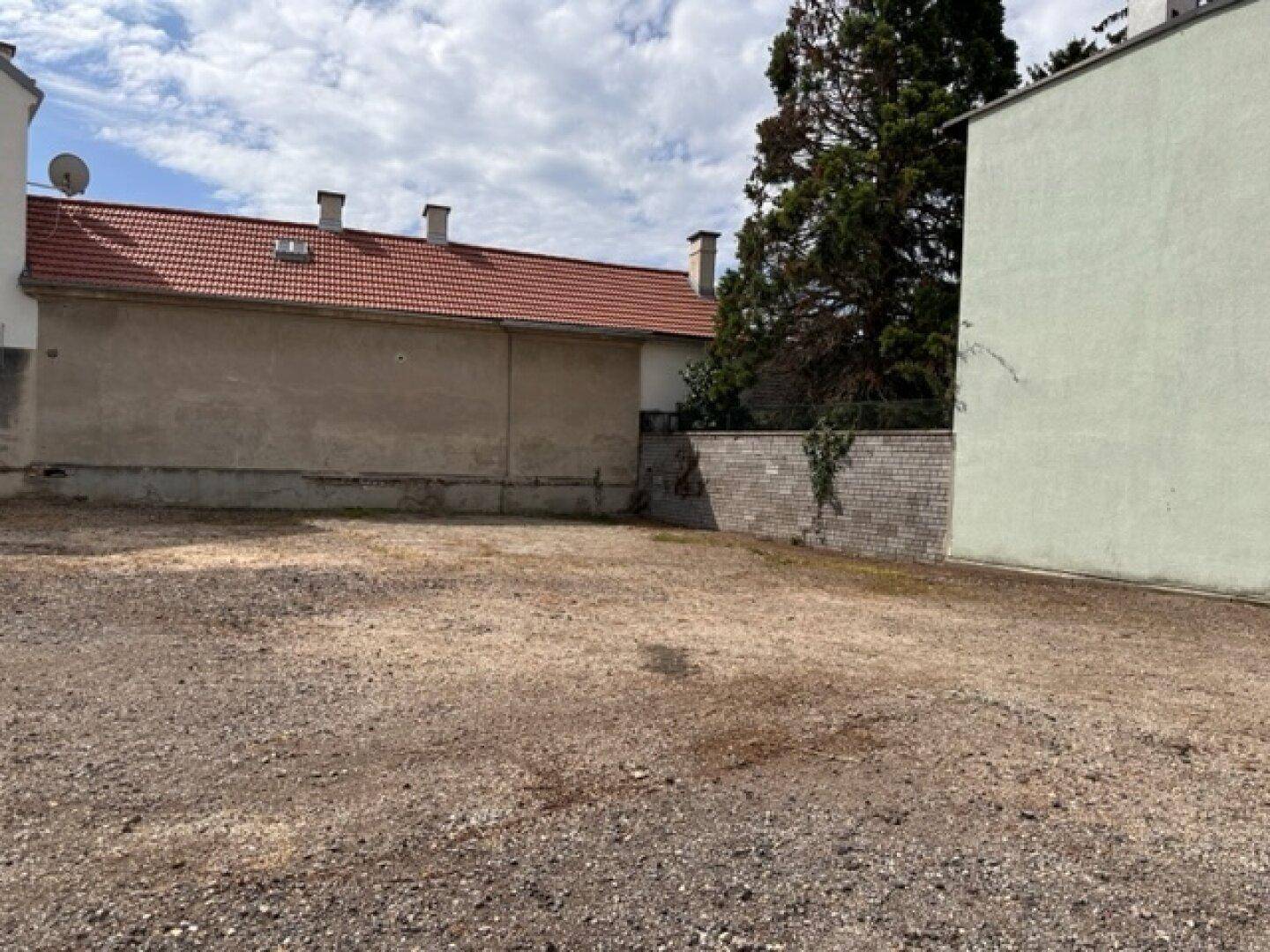 Freies Kiesgrundstück umgeben von Gebäuden, einer Ziegelmauer und einem Baum unter bewölktem Himmel.