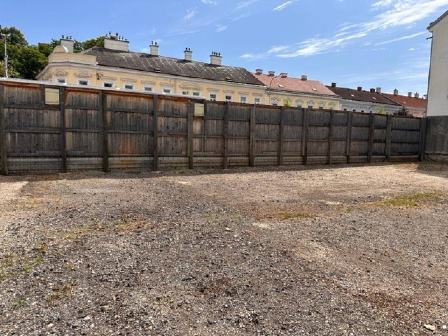 Weitläufiges Kiesgrundstück mit einem hohen Holzzaun und historischen Gebäuden im Hintergrund.