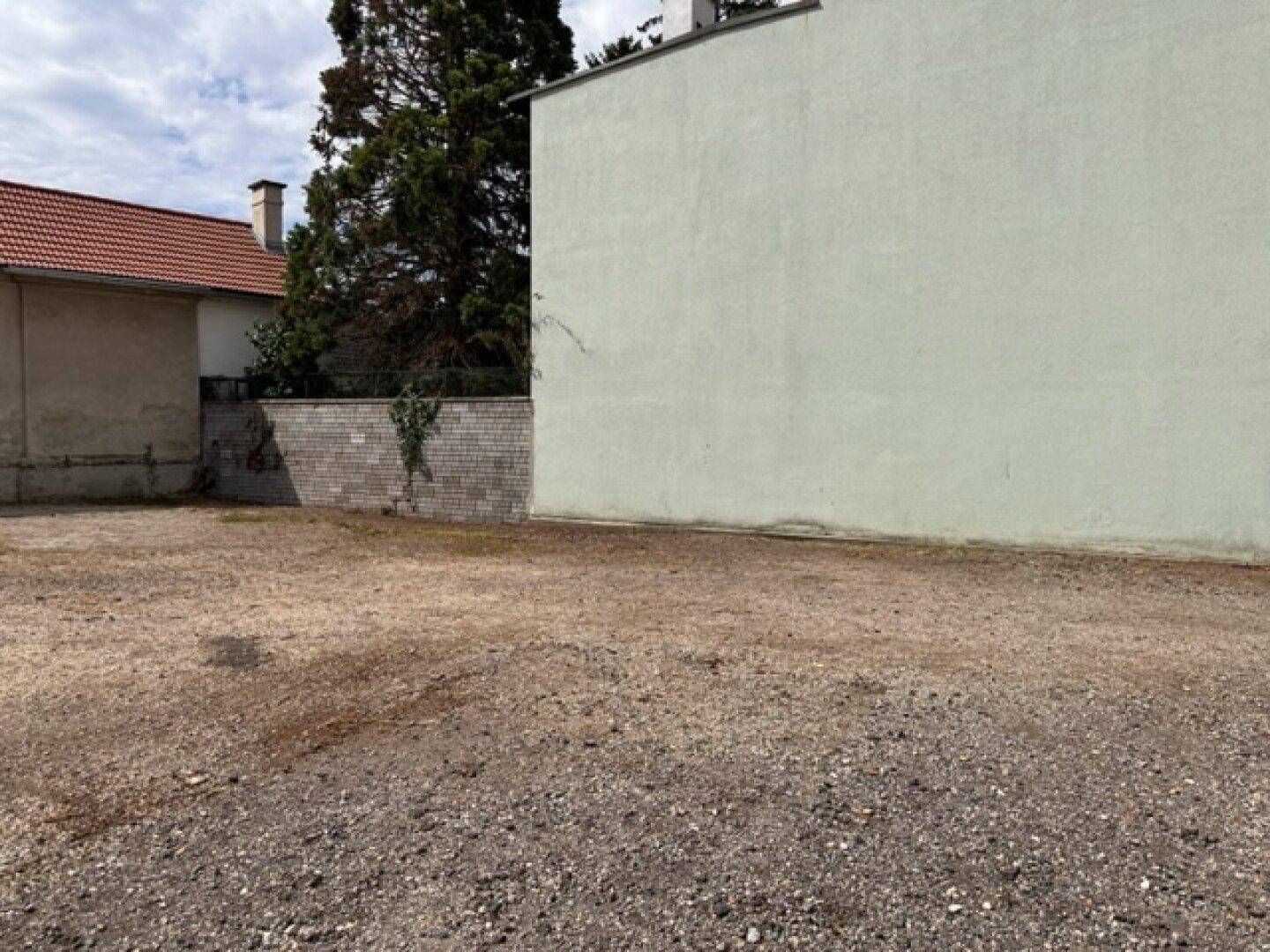 Weitläufiges Kiesgrundstück mit einer hohen Gebäudewand und einem Baum im Hintergrund.