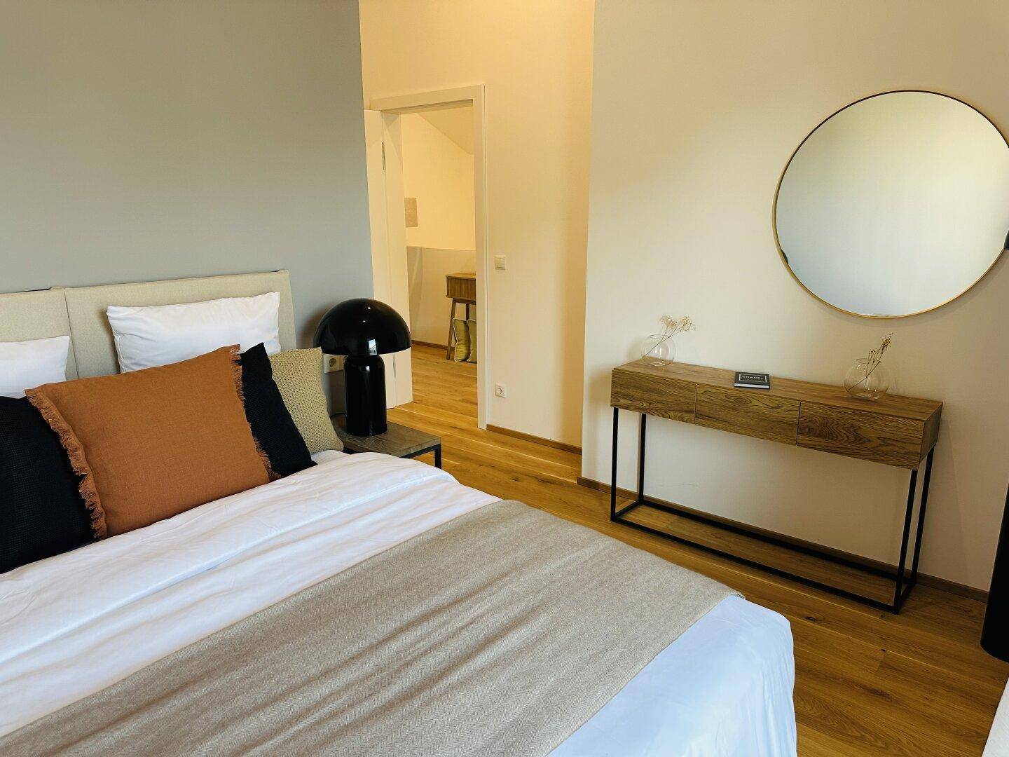 Schlafzimmer mit Holzboden, Konsole und rundem Spiegel, offen zu einem weiteren Bereich.