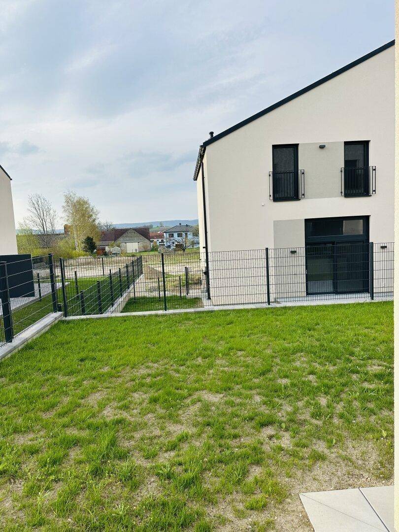 Grüner Rasenbereich neben einem modernen Neubau mit Zaun und Blick auf die ländliche Umgebung.
