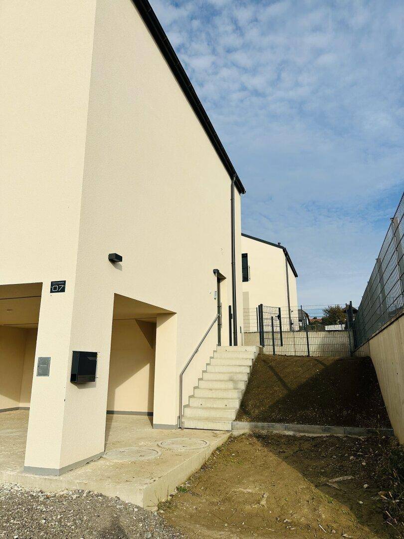 Seitenansicht eines Neubaus mit Außentreppe und Eingangsbereich unter leicht bewölktem Himmel.