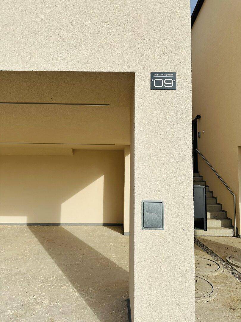 Eingangsbereich eines Neubaus mit überdachtem Stellplatz und Hausnummer an der Fassade.