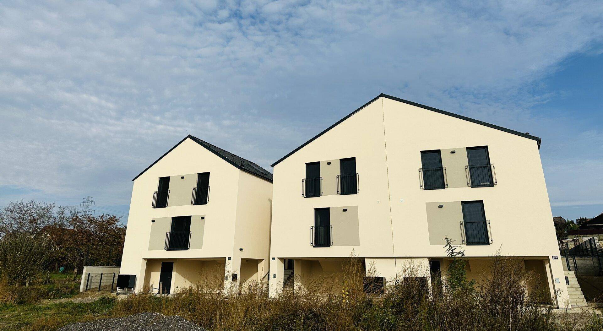 Zwei moderne Neubau-Einfamilienhäuser mit heller Fassade, Satteldach und französischen Balkonen.