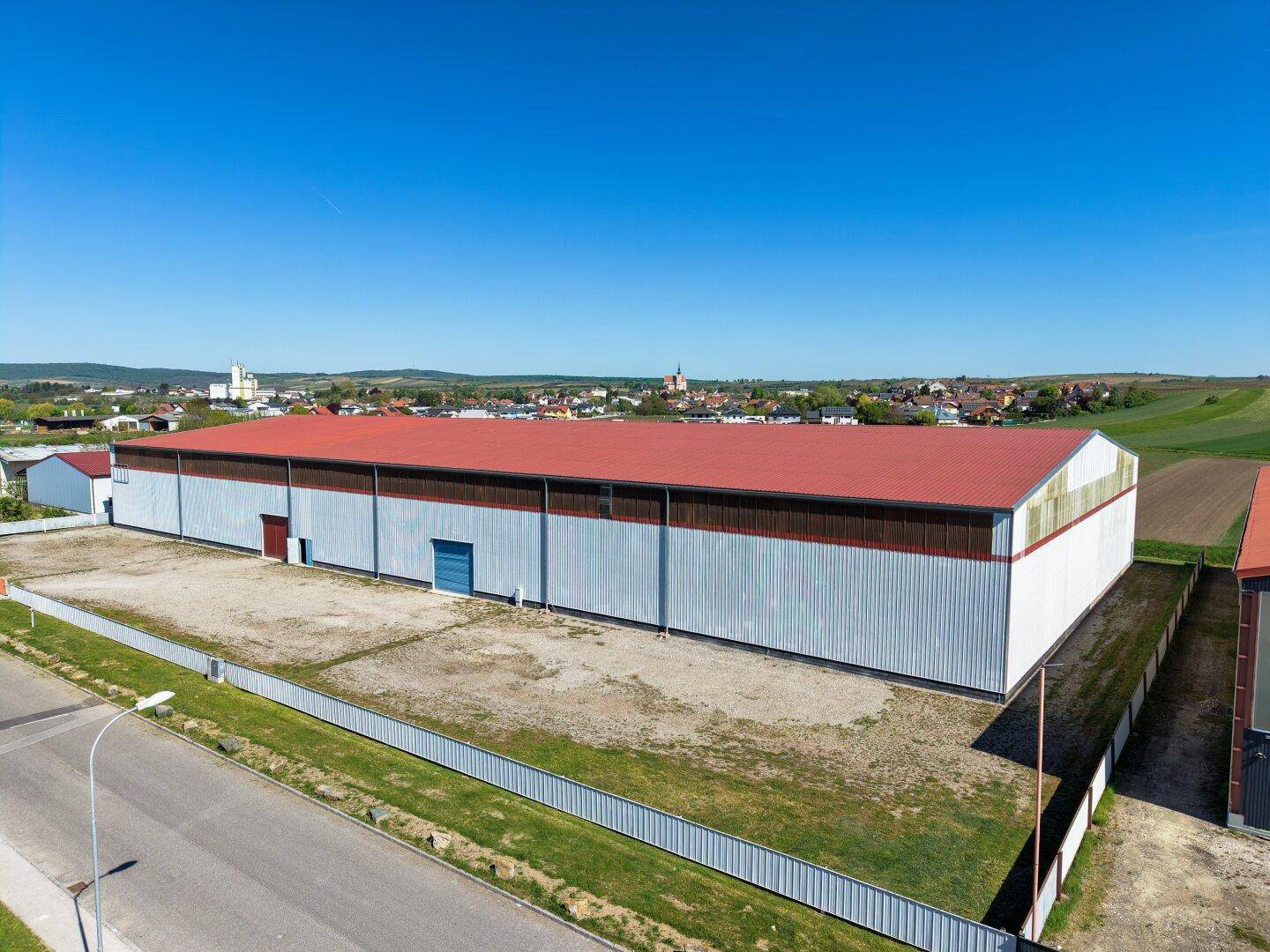 Ansicht einer langen Industriehalle mit rotem Dach und hellen Fassaden, gelegen in einer ländlichen Umgebung.