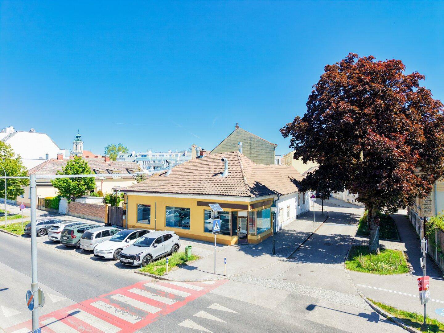 Gelbes Gebäude mit Ziegeldach an einer Straße mit parkenden Autos und einem großen Baum.