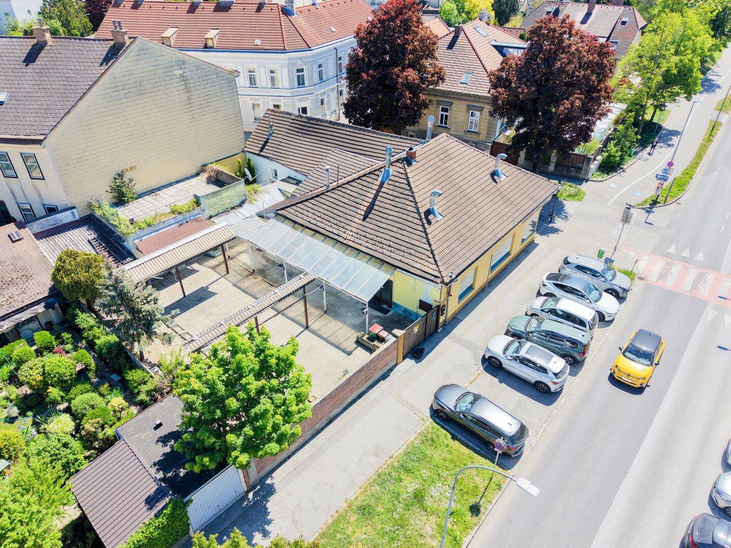 Rückansicht eines Gebäudes mit überdachtem Bereich, kleinem Garten und parkenden Fahrzeugen an der Straße.