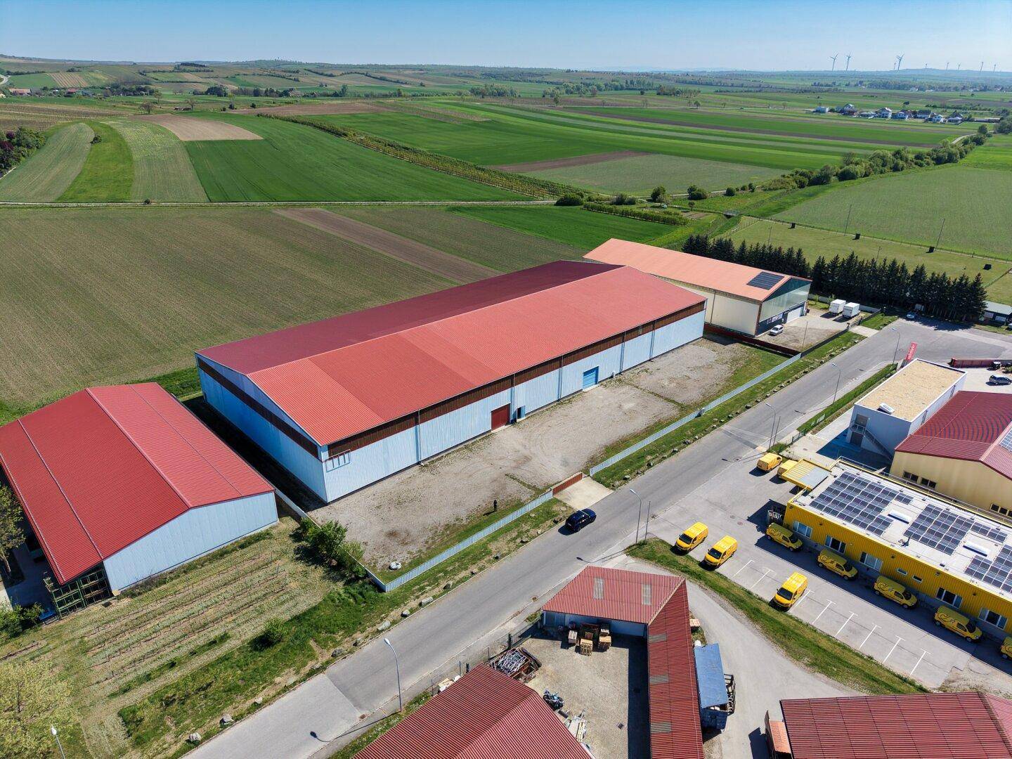 Weitläufige Luftaufnahme einer großen Lagerhalle mit rotem Dach, umgeben von grünen Feldern und Zufahrtsstraßen.