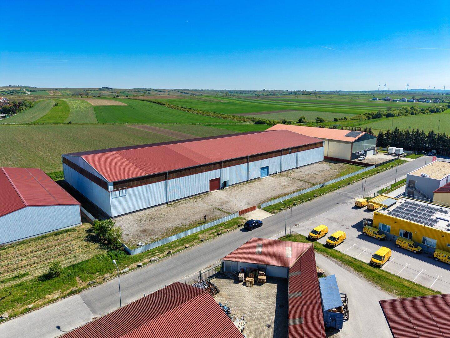 Panoramische Luftaufnahme eines Gewerbegebiets mit großer Lagerhalle, umgeben von weiten grünen Feldern.