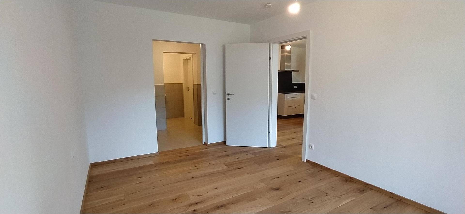 Leerer Raum mit Holzboden und Zugang zu weiteren Zimmern, darunter ein gefliestes Bad.