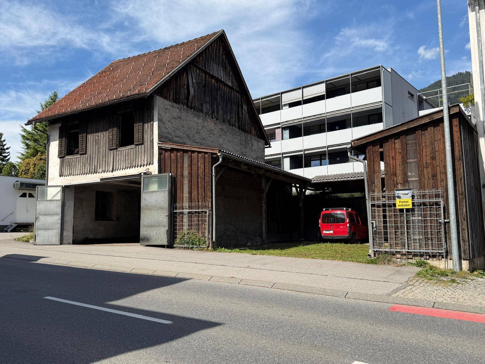 Ansicht des alten Gebäudes mit angrenzendem überdachtem Parkplatz und einem roten Fahrzeug, Berge im Hintergrund.