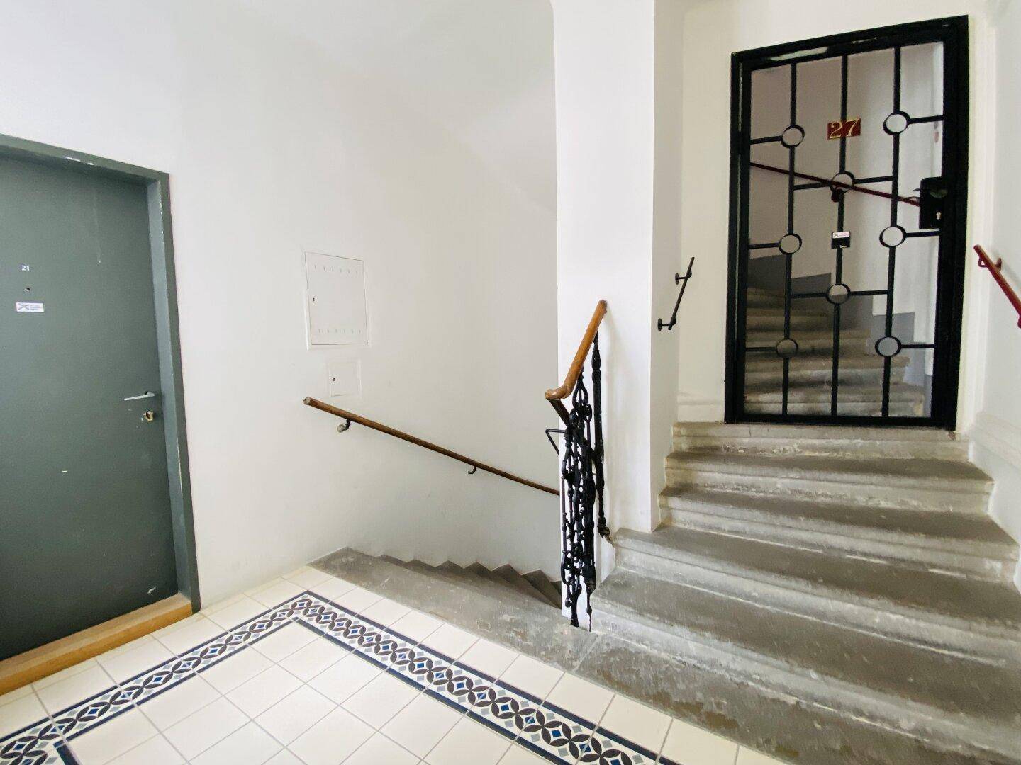 Eingangsbereich mit Treppe, schmiedeeisernem Geländer und gemusterten Bodenfliesen.