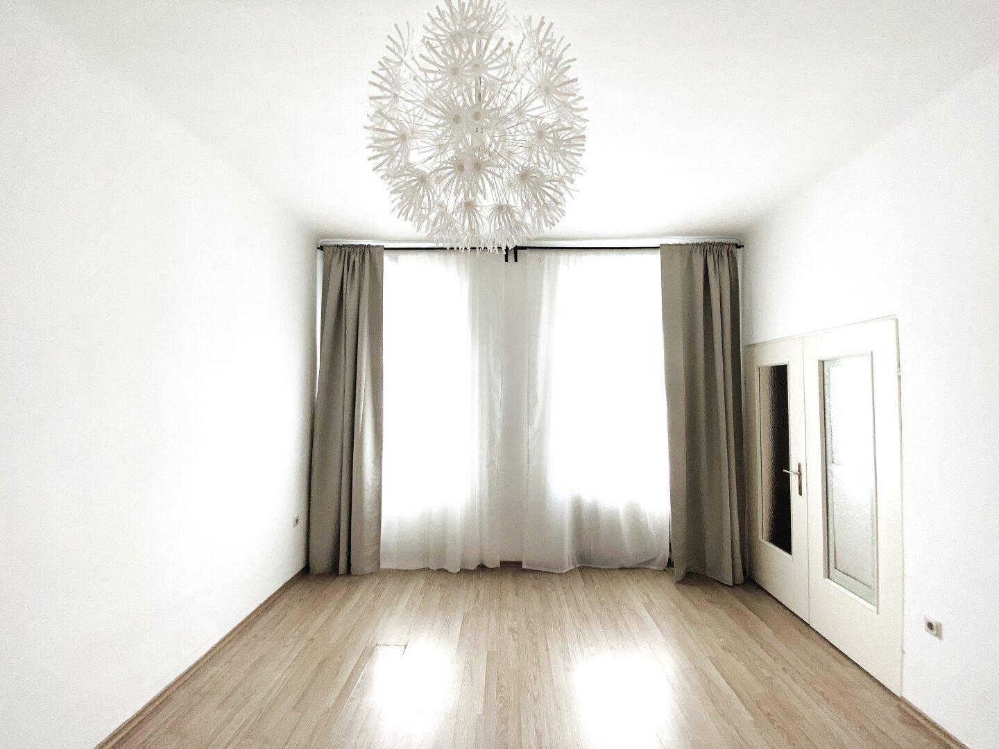 Helles Wohnzimmer mit hellem Parkettboden, großen Fenstern und einer modernen Deckenleuchte.