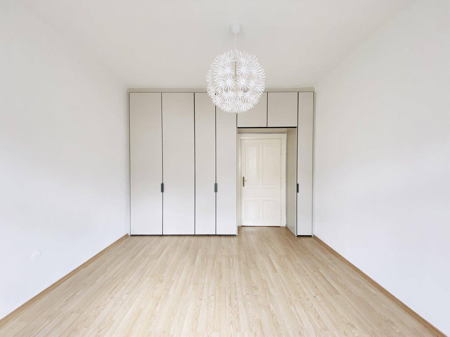 Schlafzimmer mit hellem Parkettboden, weißen Einbauschränken und einer modernen Deckenleuchte.