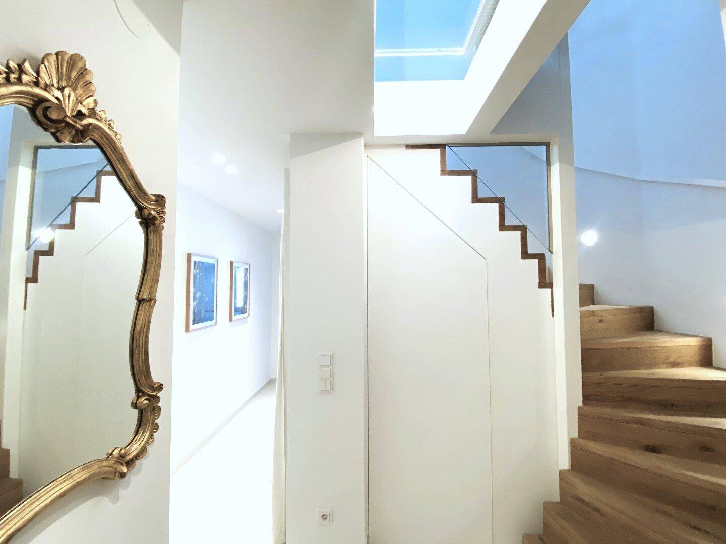 Heller Flur mit Holztreppe, modernem Glasgeländer und einem dekorativen Spiegel.