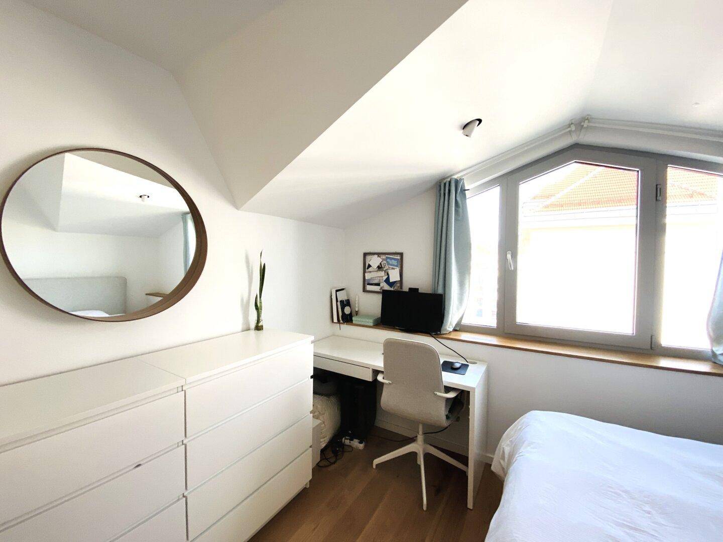 Schlafzimmer mit weißer Kommode, rundem Spiegel und Arbeitsbereich am Fenster.