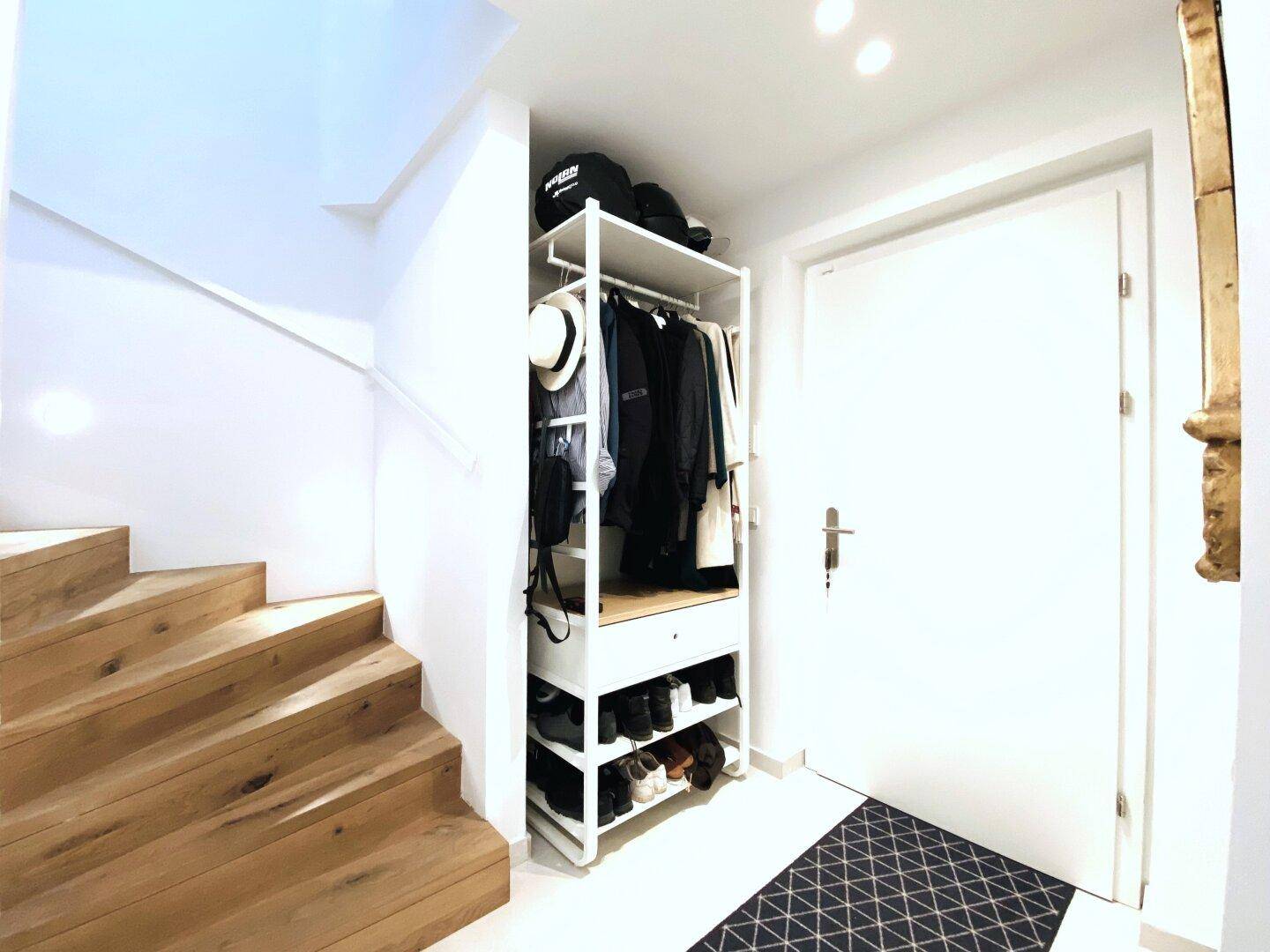 Eingangsbereich mit offener Garderobe, Holztreppe und moderner Fußmatte.