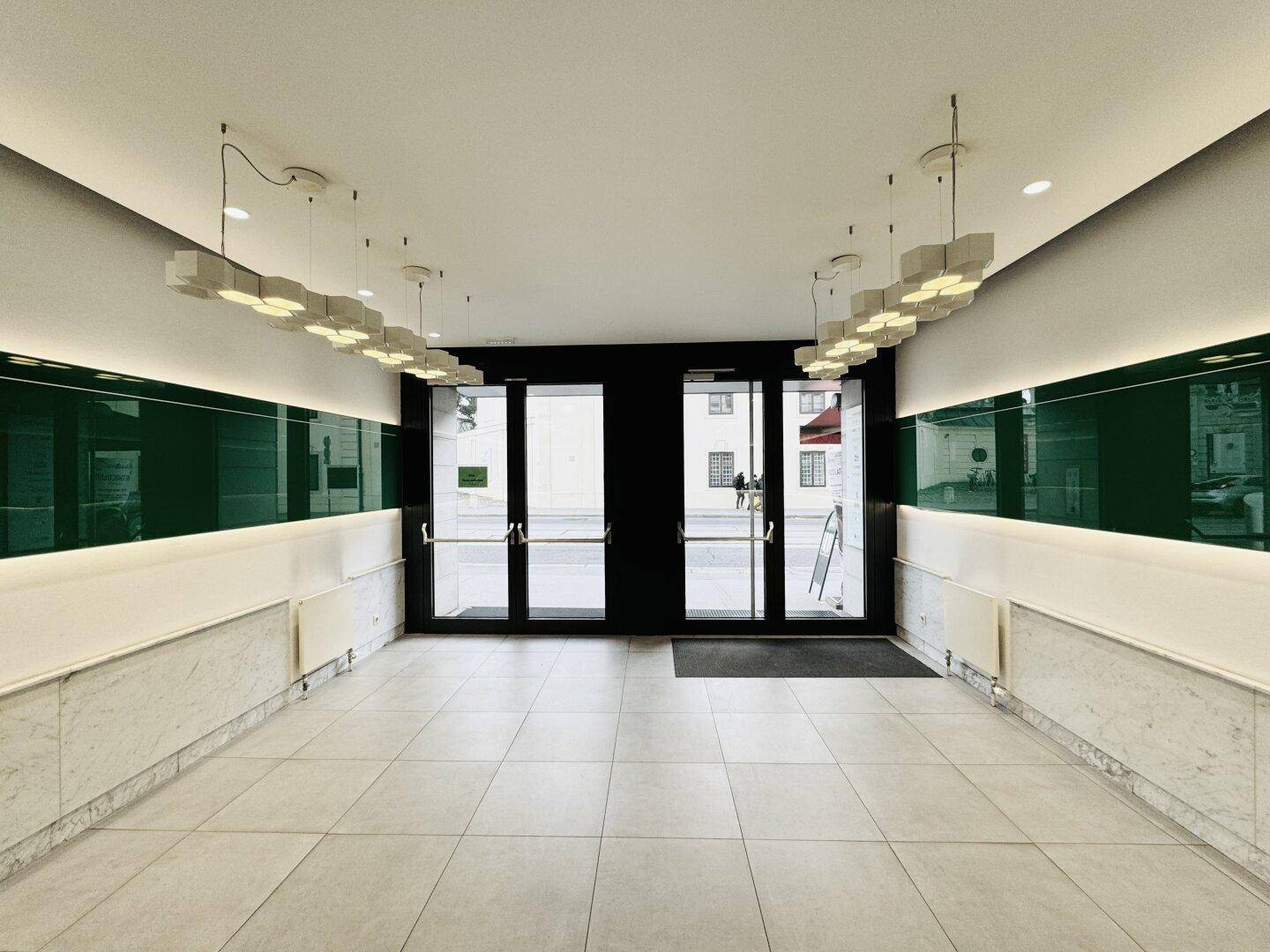 Moderner Eingangsbereich mit großen Glastüren, auffälligen Deckenleuchten und grünen Wandpaneelen.