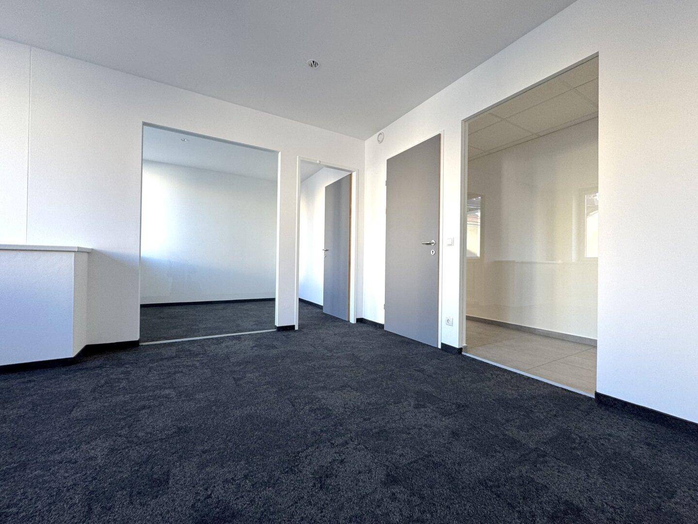 Geräumiger, leerer Bürobereich mit dunklem Teppichboden und mehreren Zugängen zu weiteren Räumen.