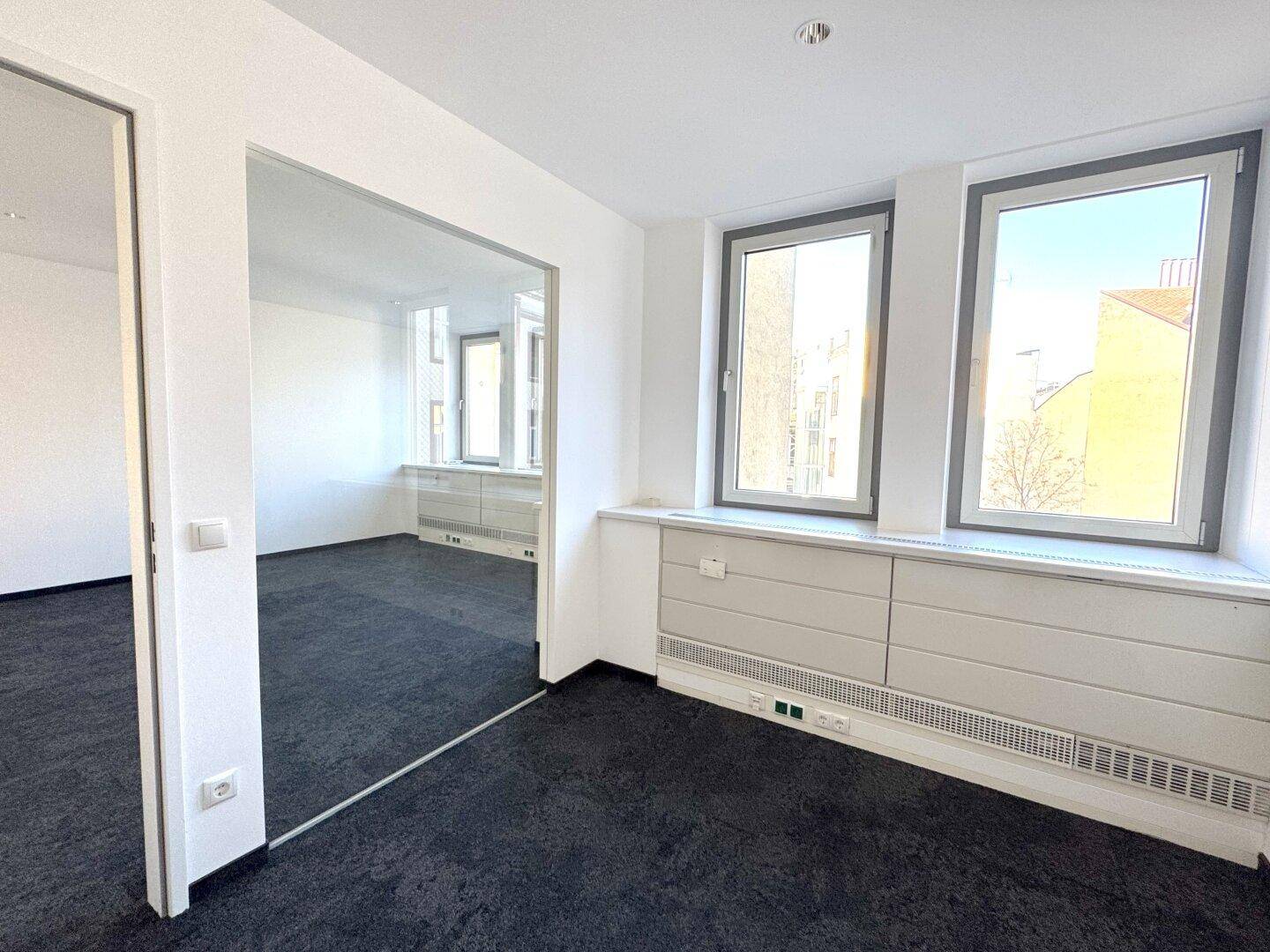 Heller Büroraum mit dunklem Teppichboden, weißen Wänden und großen Fenstern mit Blick auf die Stadt.