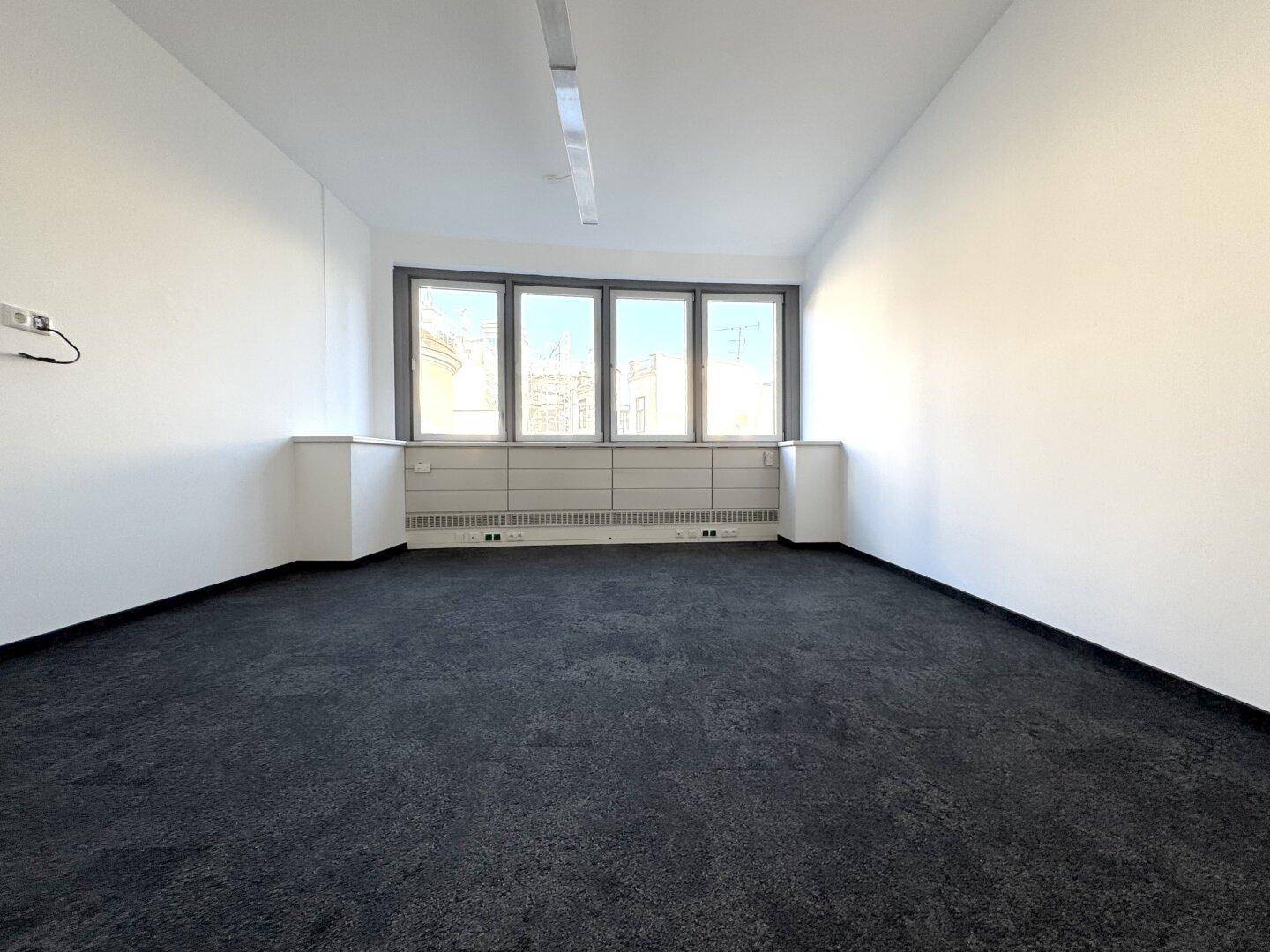 Leerer Büroraum mit dunklem Teppichboden und einer Fensterfront, die für viel Helligkeit sorgt.