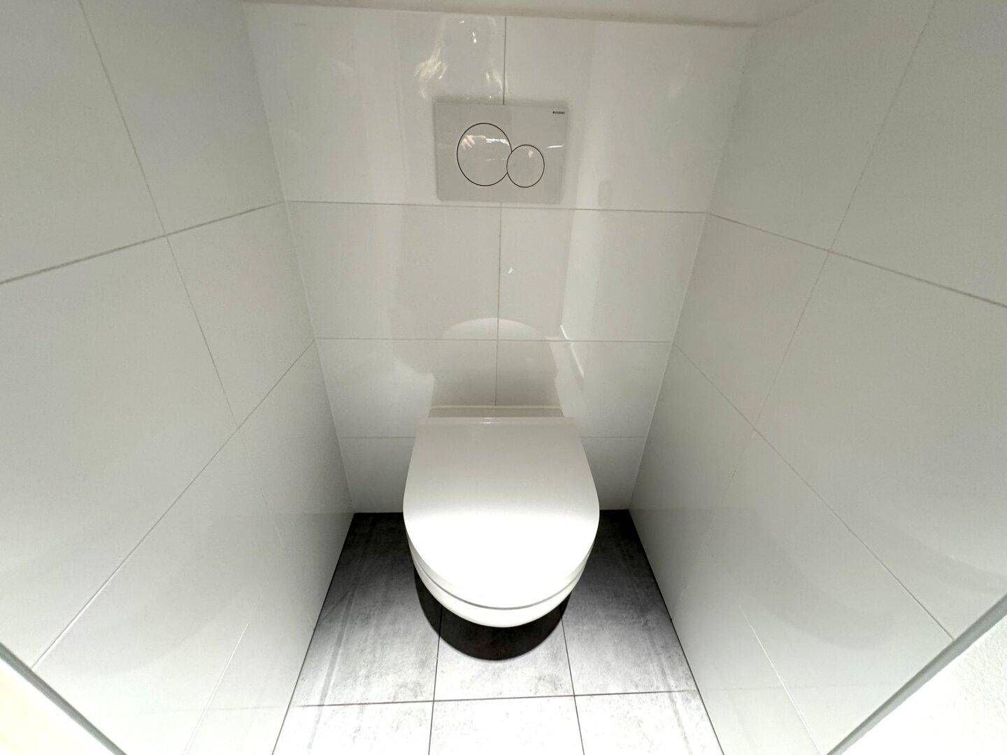 Sauberer Toilettenraum mit wandhängendem WC, weißen Fliesen an den Wänden und dunklem Fliesenboden.