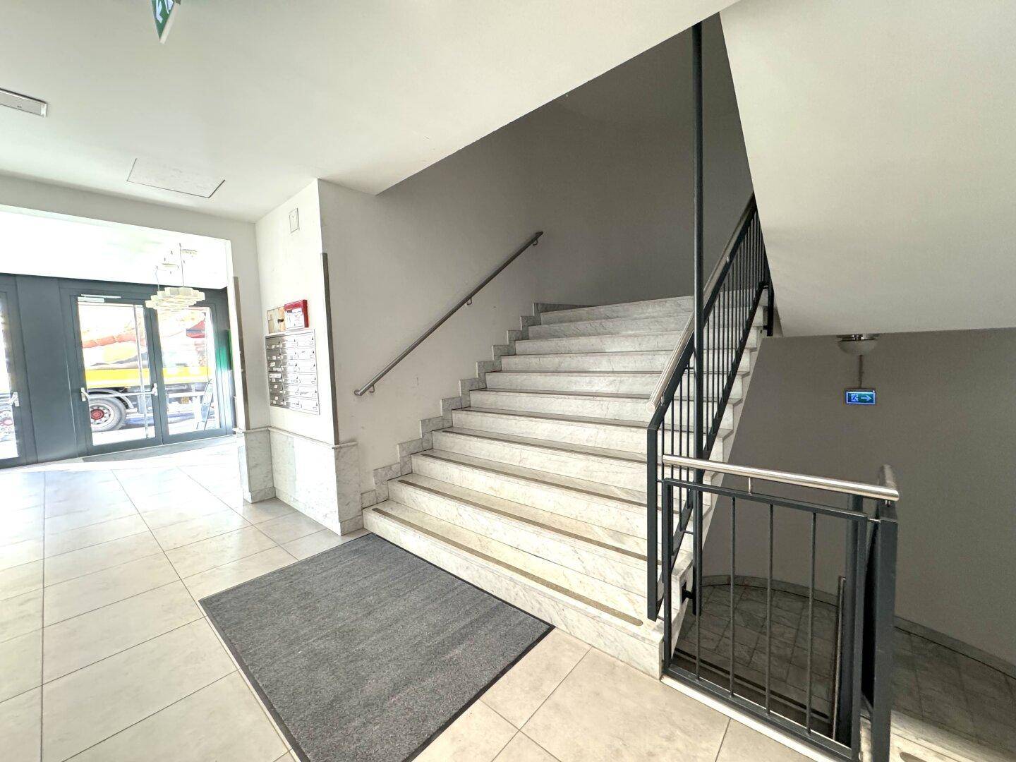 Heller Eingangsbereich mit breiter Marmortreppe, Metallgeländer und großen Glastüren zum Außenbereich.
