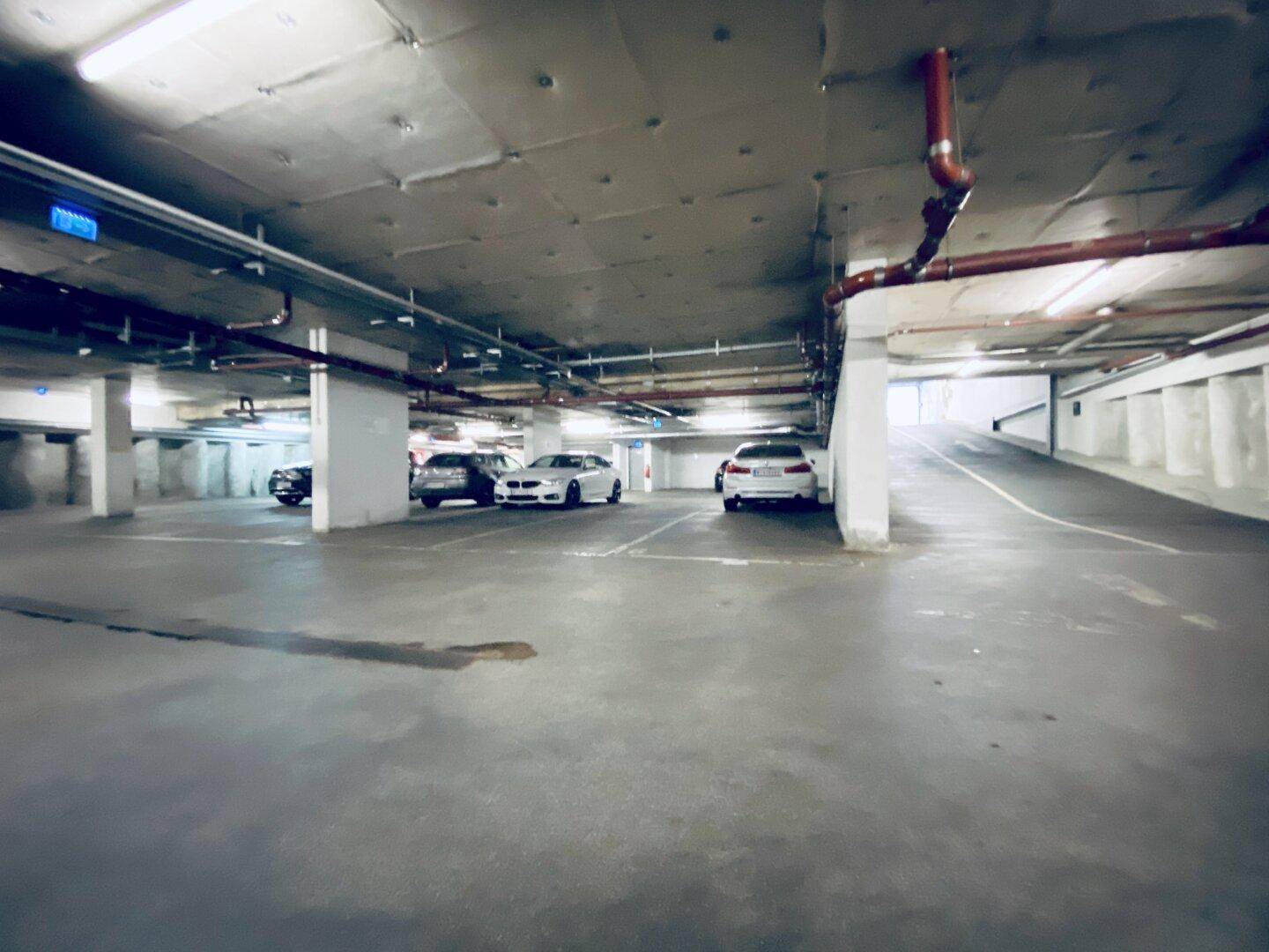 Geräumige Tiefgarage mit mehreren geparkten Fahrzeugen und sichtbaren Versorgungsleitungen an der Decke.