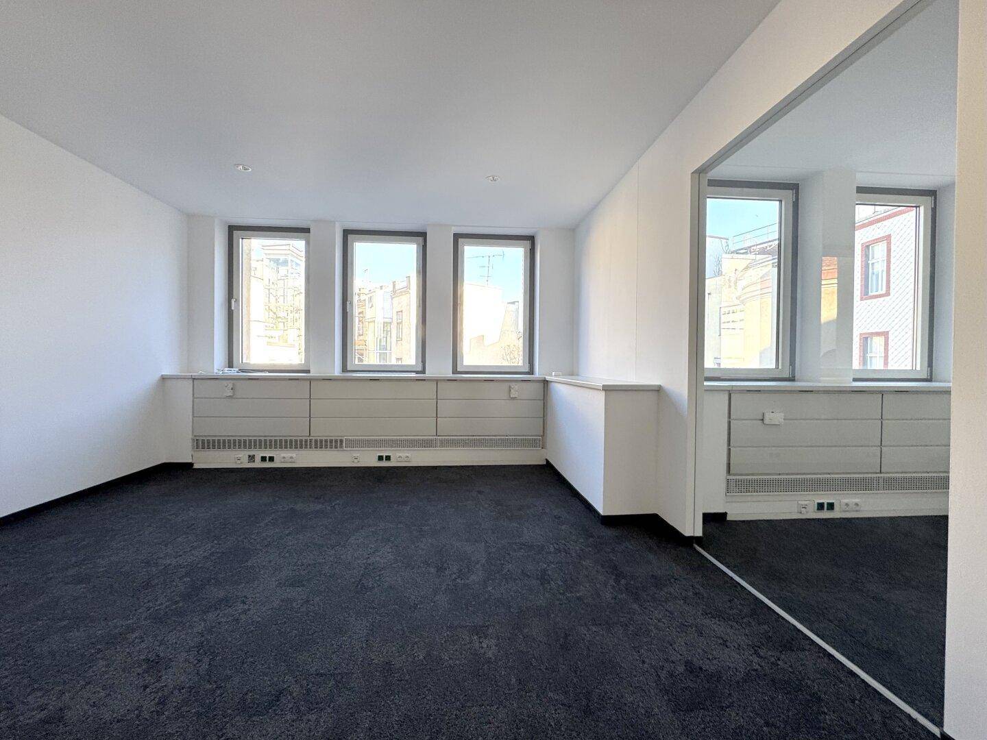 Heller Büroraum mit dunklem Teppichboden, weißen Wänden und drei Fenstern mit Blick auf die Umgebung.
