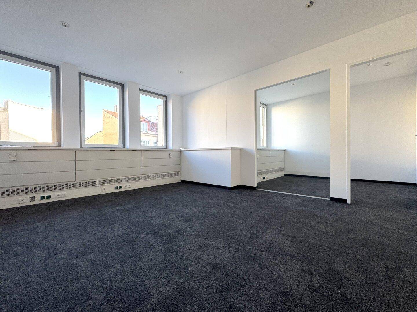 Geräumiger Büroraum mit dunklem Teppichboden, weißen Wänden und Fenstern, die viel Tageslicht hereinlassen.
