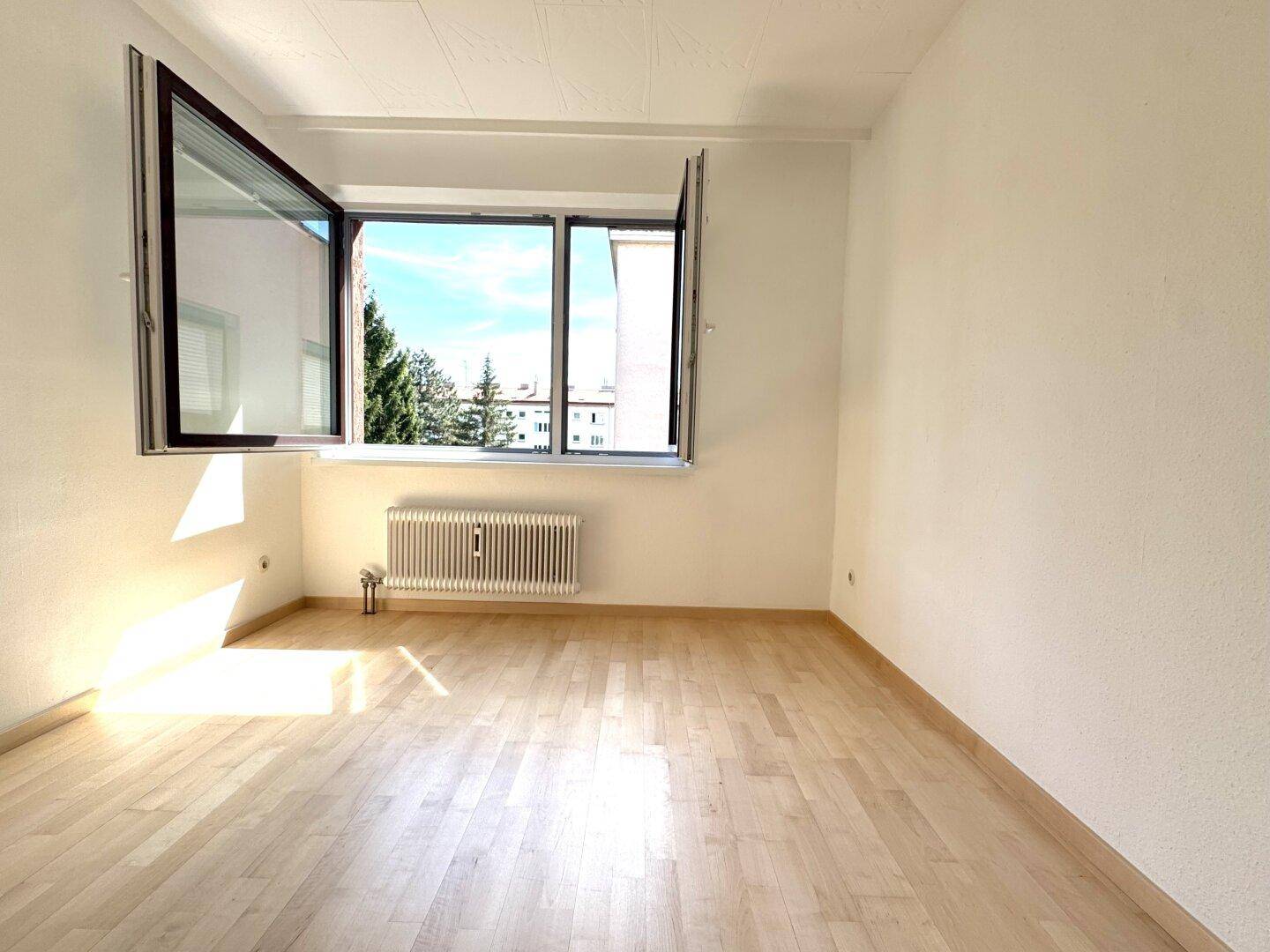 Helles, leeres Zimmer mit hellem Holzboden, weißen Wänden und einem Fenster mit Ausblick.