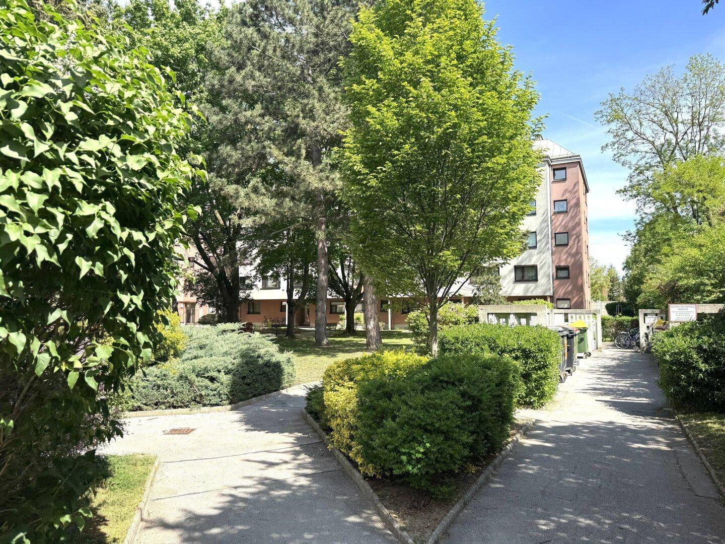 Grüner Außenbereich mit Gehwegen, Büschen und Wohngebäuden im Hintergrund an einem sonnigen Tag.