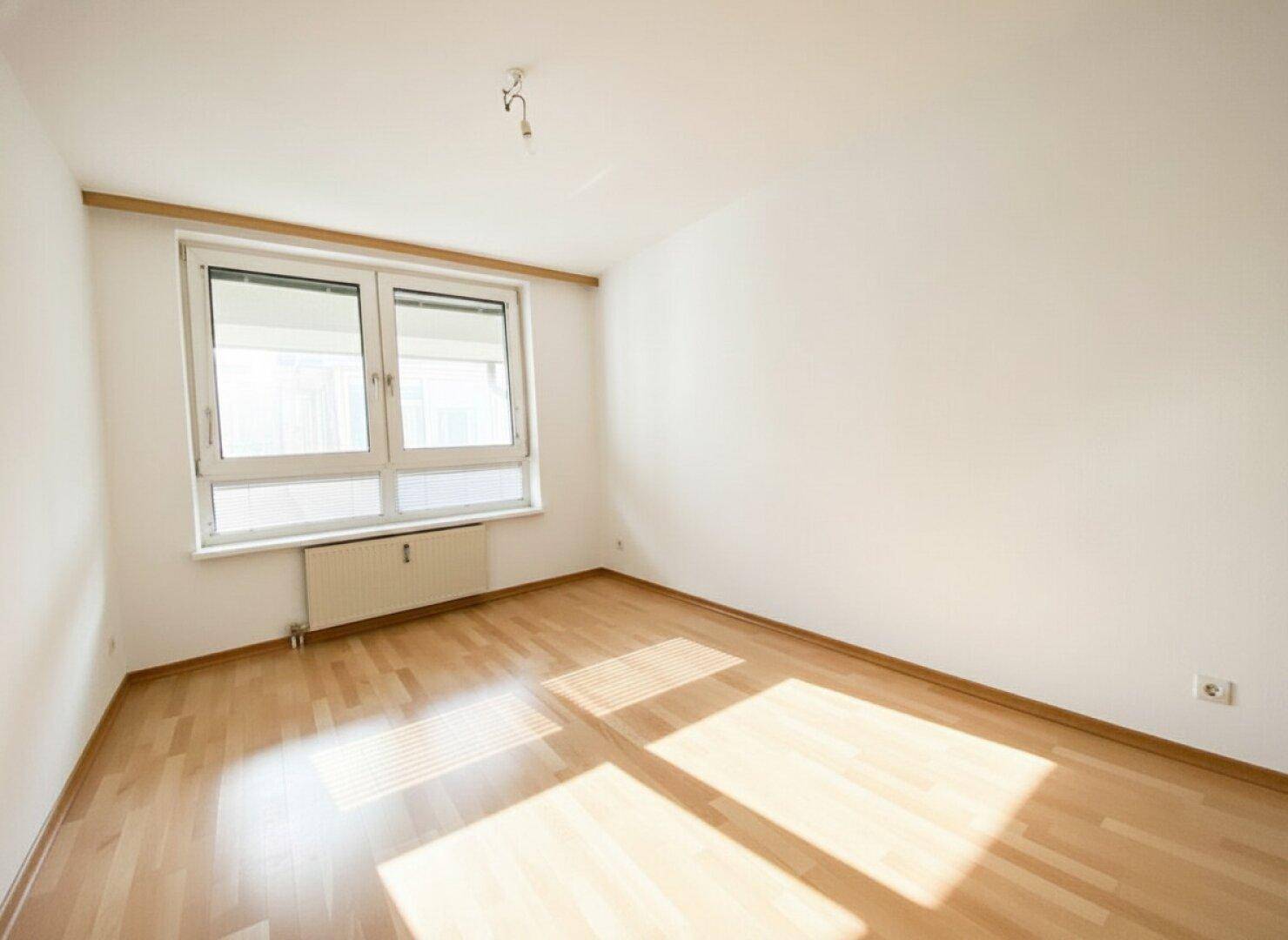 Helles, leeres Zimmer mit Holzboden und großem Fenster, ideal als Schlaf- oder Arbeitszimmer.