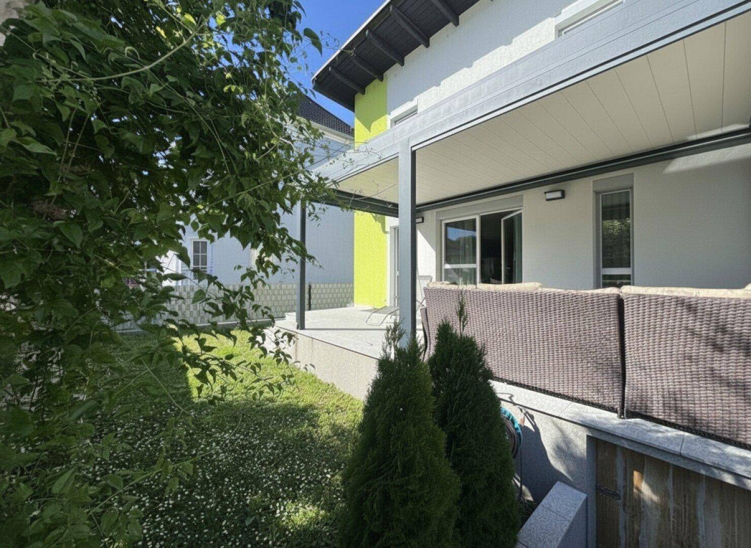 Überdachte Terrasse mit Lounge-Möbeln und Blick in den grünen, sonnigen Gartenbereich.