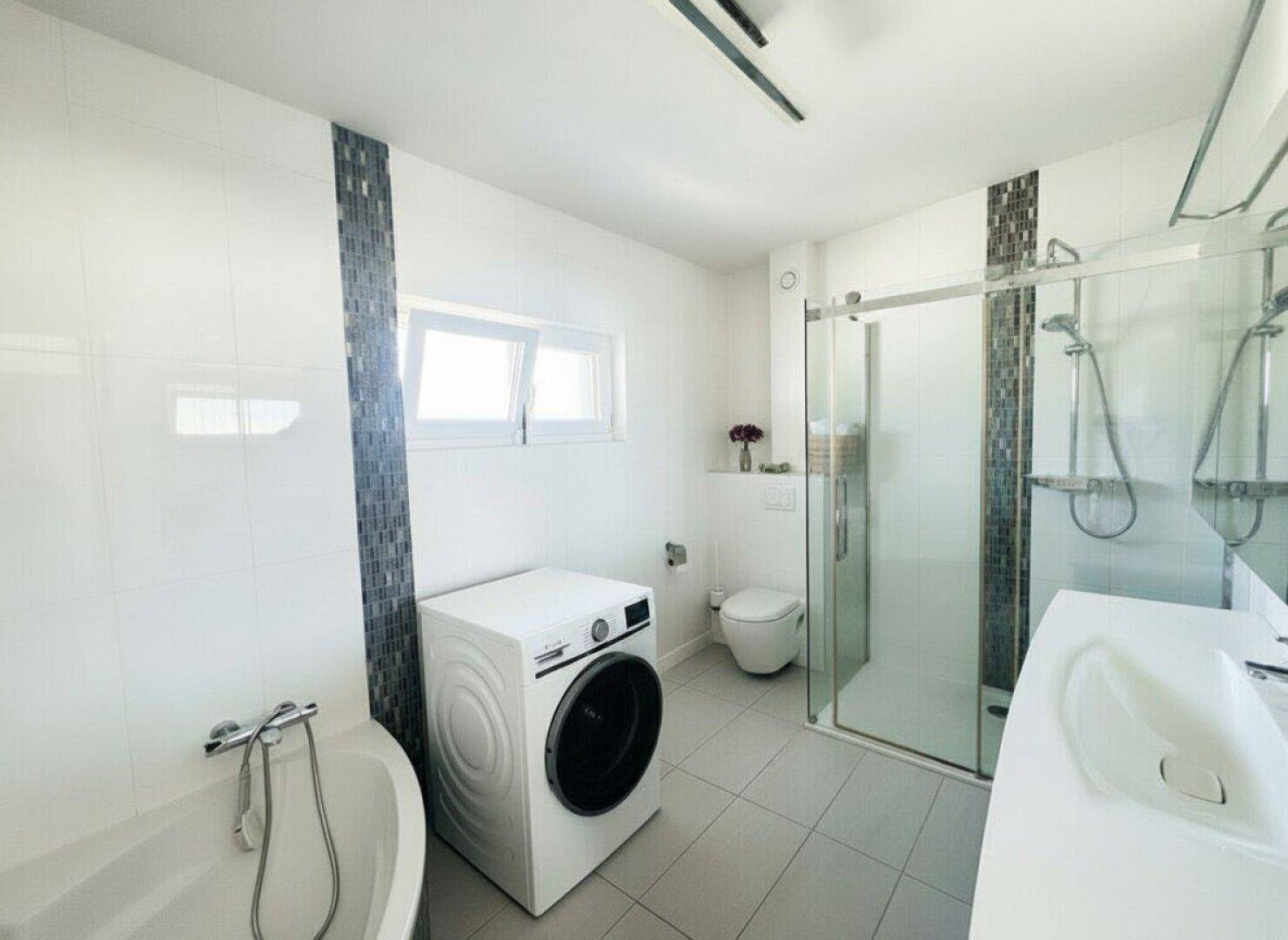 Helles Badezimmer mit Dusche, Badewanne, Waschmaschine und Fenster für Tageslicht.